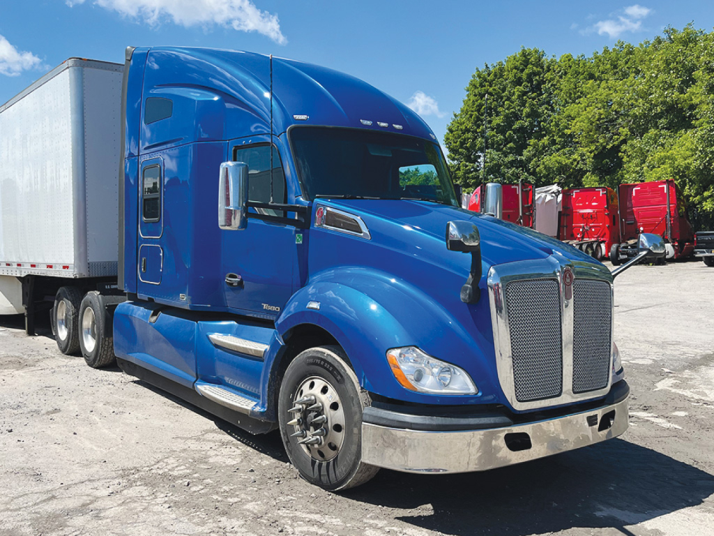 Kenworth T680 blue sleeper cab tractor-trailer in a salvage lot, front three-quarter view, with chrome grille, exposed wheels, and a white trailer.