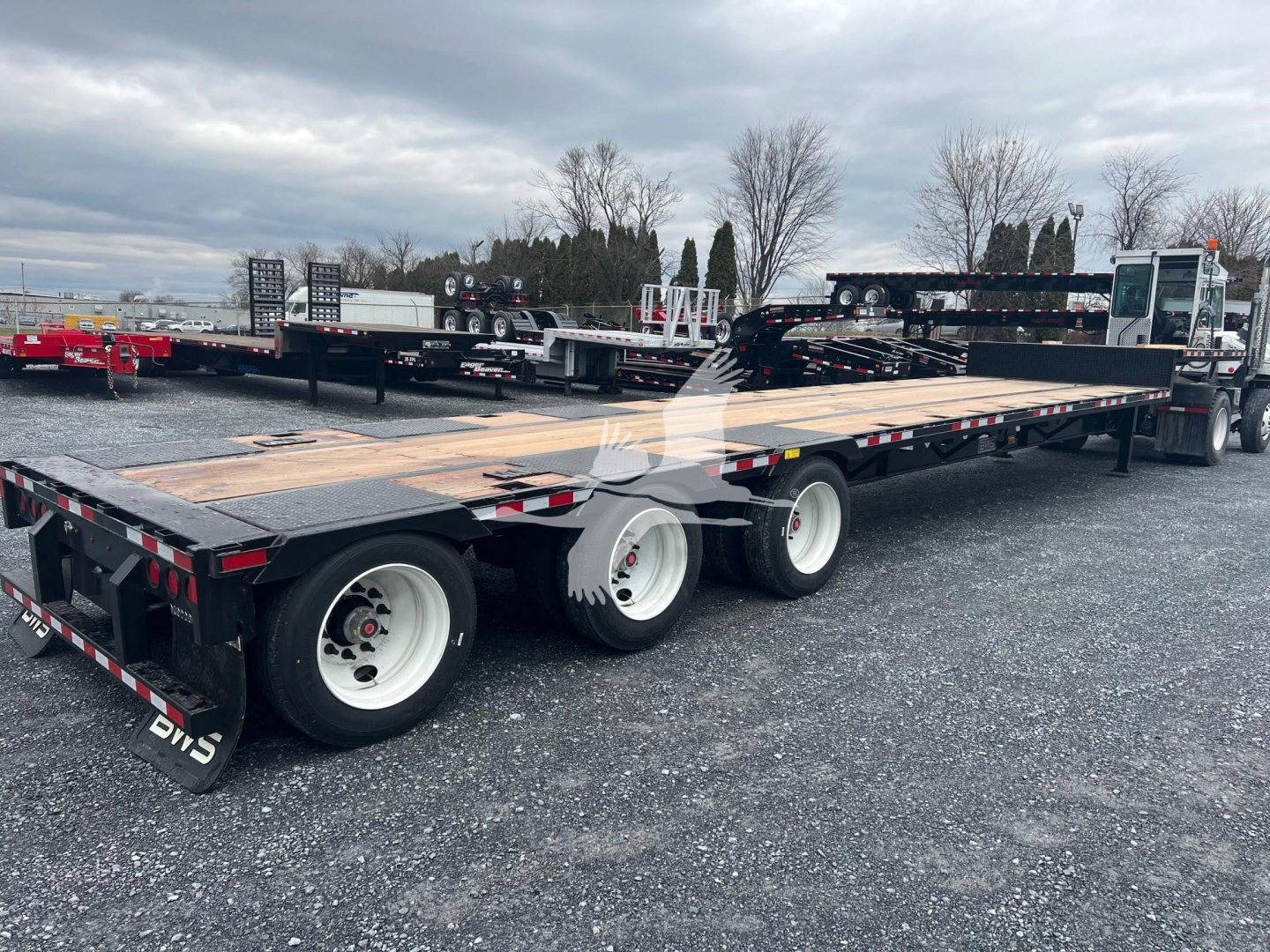 Trailer showing a 3-axle drop deck middle lift with wood deck and white wheels, rear view with flatbed sections and equipment in background.