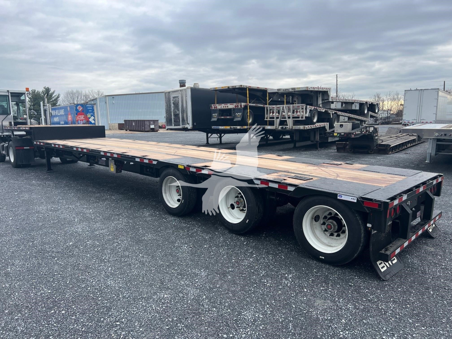 Mid-length view of a drop deck trailer with three axles and wooden deck, parked in lot with other equipment in background.