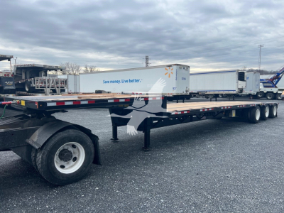 Trailer with three-axle middle lift, long drop deck, white and blue branding visible on side panels, stock trailer in lot with other trailers in background.
