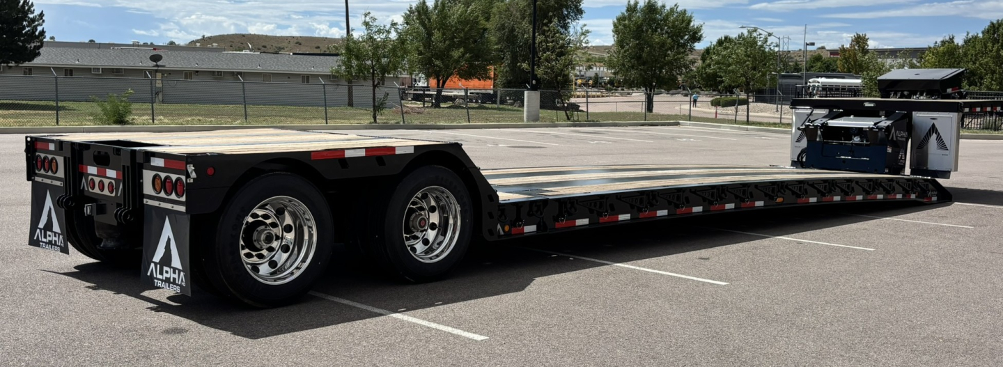 Lowboy trailer with triple rear axles and extended flatbed. Brand logos on rear include Alpha and warning triangles. Flatbed surface shows no visible writings.
