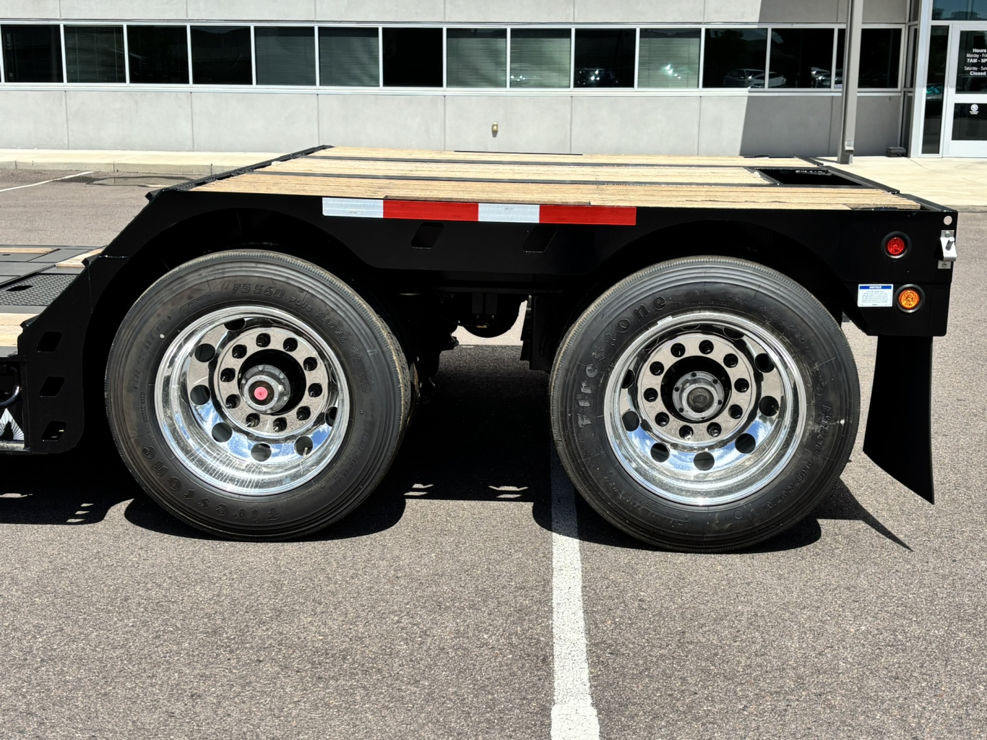 Trailer side view showing dual axles and wood deck, black frame, chrome wheels, reflective tape, and small rear fender area.