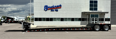 Lowboy trailer with triple-axle rear, long extendable detachable gooseneck in front, black deck with red and white safety markings, parked in dealer lot.