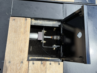 Trailer component inside a lowboy deck with hydraulic detachment mechanism and extendable double drop; visible hydraulic ram, mounting brackets, and black painted metal housing.
