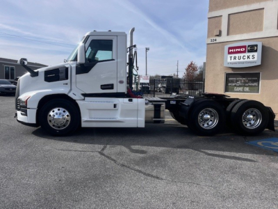 Truck cab and chassis of a 2026 Mack PR64T conventional daycab, side view with white paint, chrome wheels, and dual rear axles, dealership building in background.
