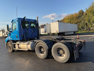 Freightliner Cascadia Daycab tractor chassis with blue cab, three axles, exposed fuel tanks, parked in lot.