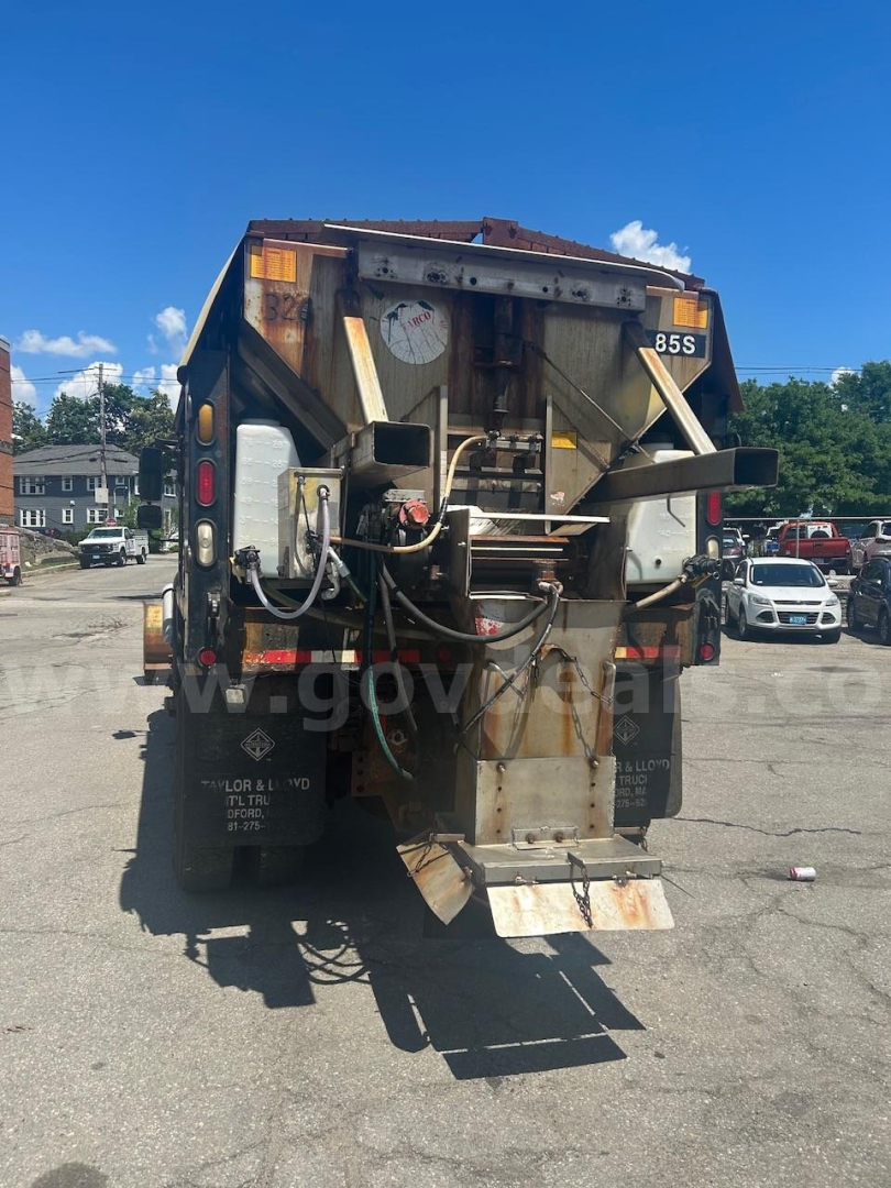 Dump truck rear showing hydraulic lines, dump bed mechanism, and tailgate with warning panels and rear lights.
