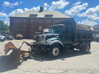 Dump truck with front plow on a clear day; green International 7400 with yellow wheels parked in a lot in front of a brick building.