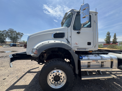 Truck cab and chassis section of a 2027 Mack GR44B showing front wheel, chrome fuel tanks, and side mirror.