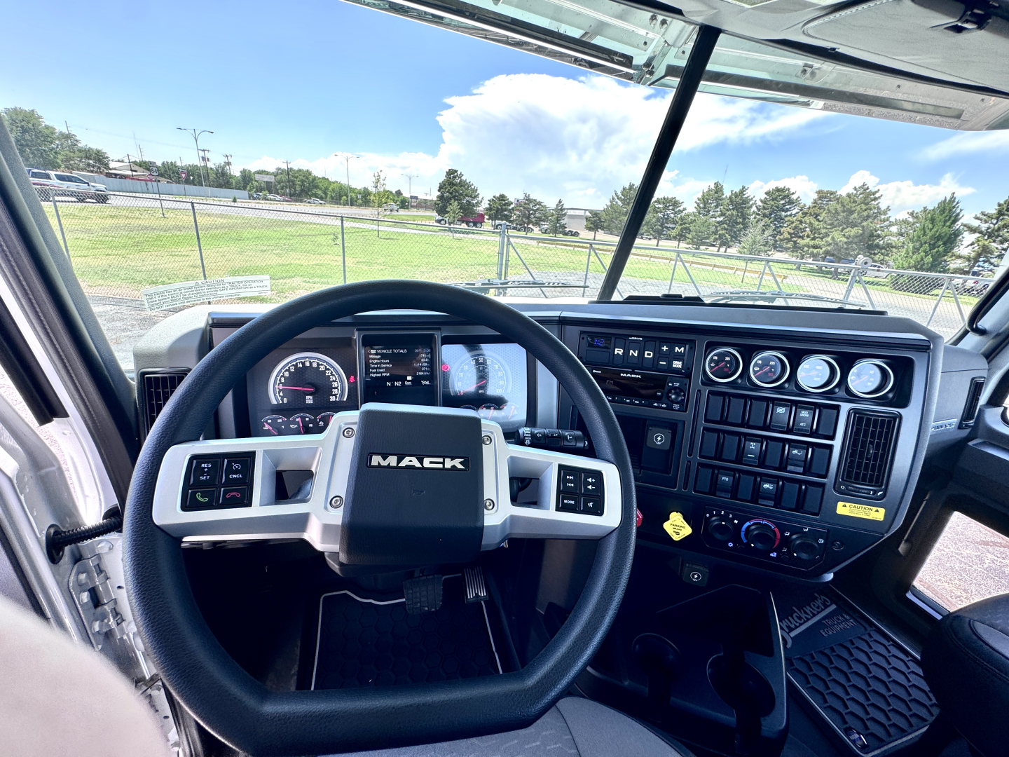 Truck cab interior showing Mack steering wheel and instrument cluster, dashboard gauges, switches, and controls; windshield reveals outside lot.