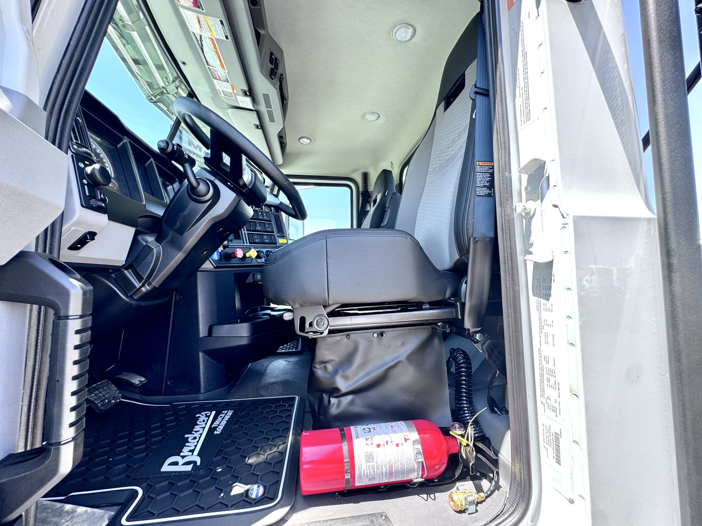 Truck cab interior of a 2027 Mack PI64T showing steering wheel, driver's seat, dashboard, and a red fire extinguisher near the door sill.
