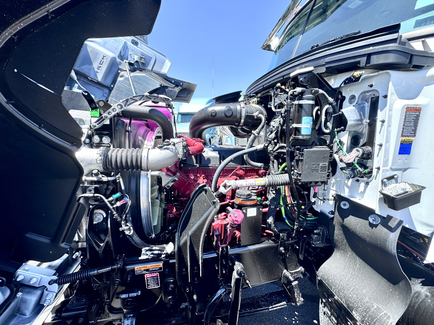 Engine bay of a new Mack PI64T with visible engine components, hoses, and wiring harnesses; includes label stickers and intake piping.