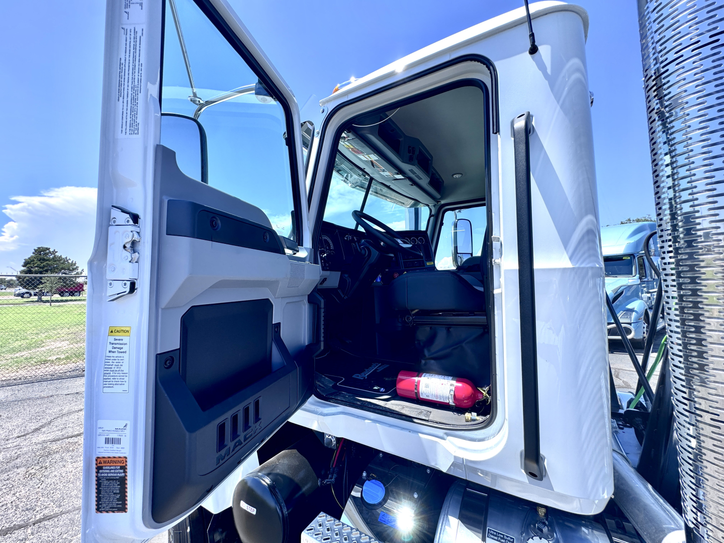 Cab interior of a 2027 Mack PI64T truck with door open showing steering wheel, seating, and dashboard; exterior white paint and chrome exhaust stack visible.