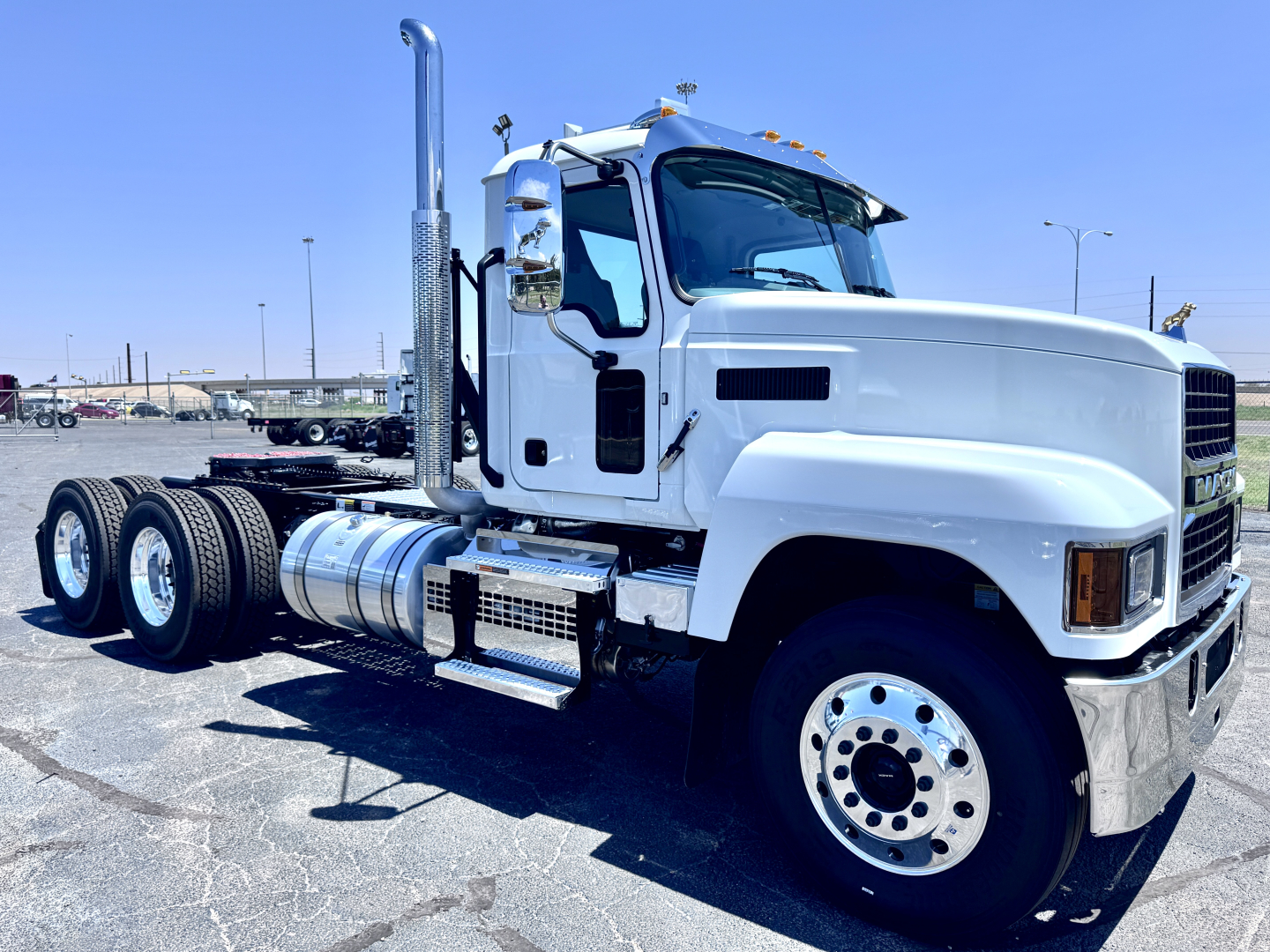 Truck cab side view showing Mack PI64T chassis and front wheel with dual rear axles, fuel tanks, and exhaust stack.