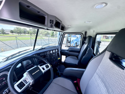Inside cab of a 2027 Mack PI64T truck showing driver's seat and passenger seat, dashboard with gauges and control panels, door, and overhead storage compartments.