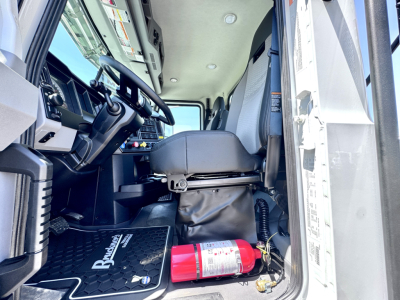 Truck cab interior of a 2027 Mack PI64T showing steering wheel, driver's seat, dashboard, and a red fire extinguisher near the door sill.