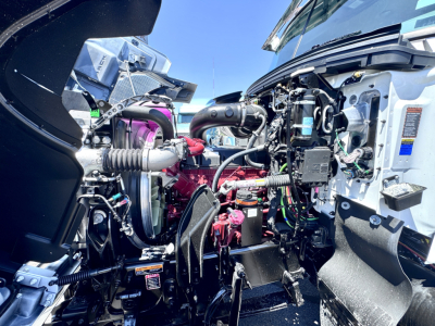 Engine bay of a new Mack PI64T with visible engine components, hoses, and wiring harnesses; includes label stickers and intake piping.