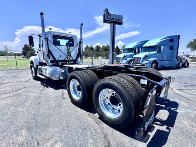 Truck chassis with dual rear axles and empty flatbed area, cab visible on left, Mack dealership sign in background