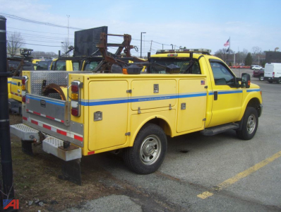 Ford F-350 Super Duty utility truck with plow, yellow body, blue stripe, service compartments, rear towing equipment, side ladder/step, parked in lot.