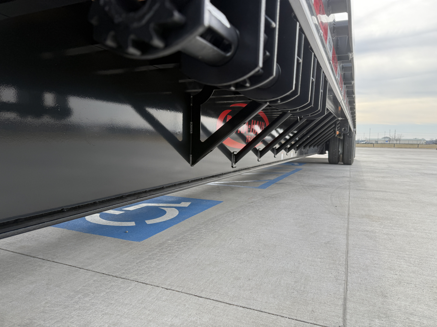 Trailer underside with hydraulic components and black steel frame, view from ground level near a blue handicapped parking space marking.
