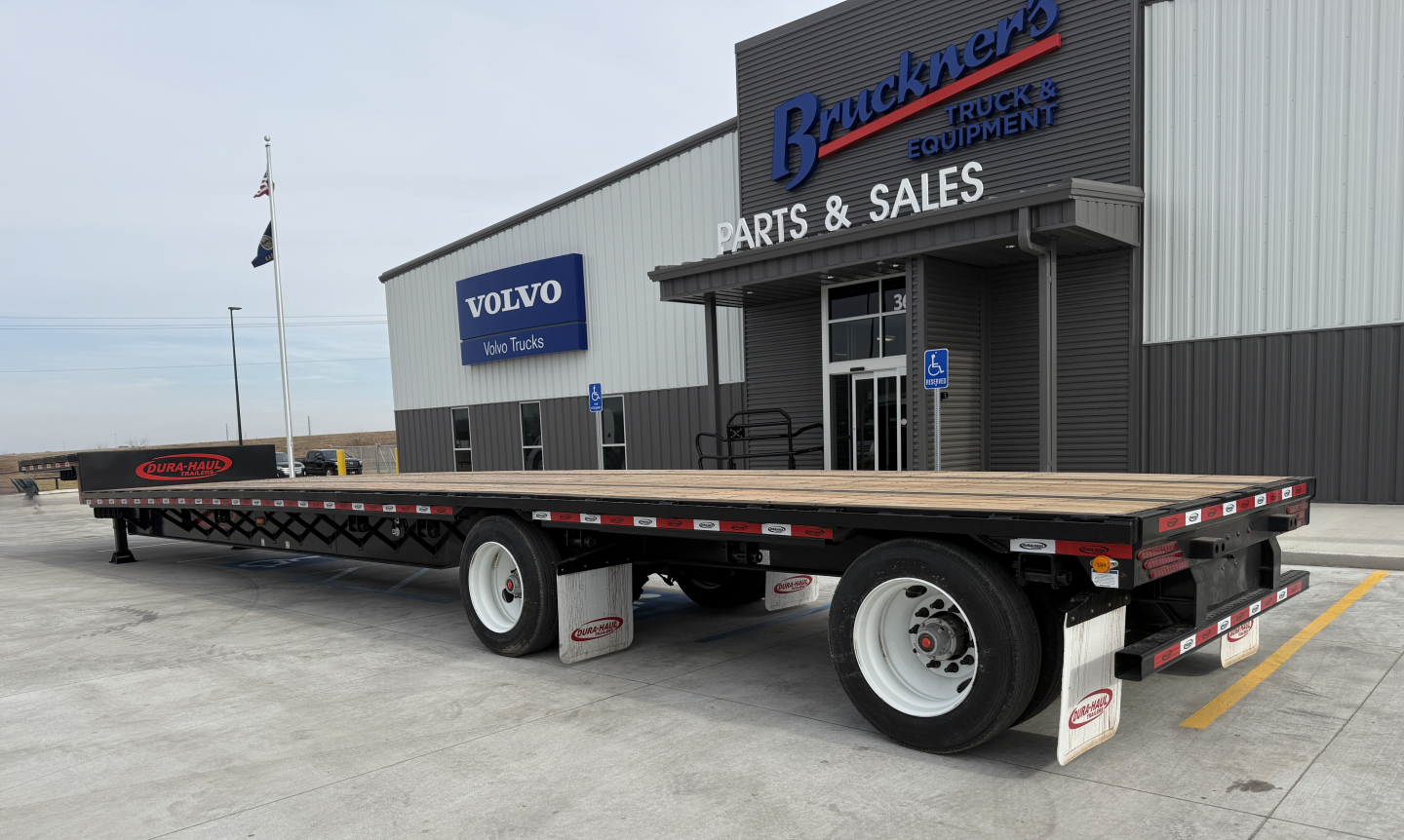 Flatbed trailer with wooden deck and dual rear axles in front of a parts and sales building. Visible text includes 'BRUCKNER'S TRUCK & EQUIPMENT' on the building, 'VOLVO' sign, and truck yard surroundings.
