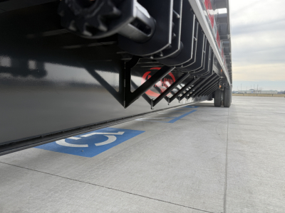 Trailer underside with hydraulic components and black steel frame, view from ground level near a blue handicapped parking space marking.