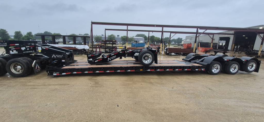Trailer deck of a lowboy trailer with multiple axles and extendable neck, black with red/white reflective stripes, brand name NMC on frame, in yard with other trailers.