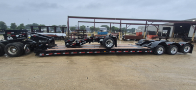 Trailer deck of a lowboy trailer with multiple axles and extendable neck, black with red/white reflective stripes, brand name NMC on frame, in yard with other trailers.