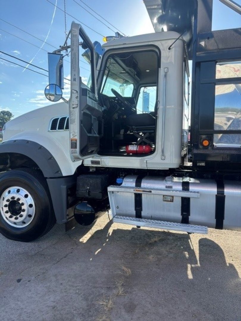 Truck cab interior visible through open driver door; front wheel, fuel tank, and ladder rack of a white 2022 Mack Granite dump truck.