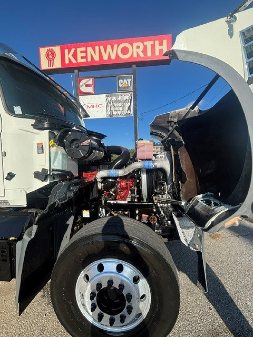 Truck engine bay of a 2022 Mack GRANITE dump truck showing engine with exposed components and chassis; visible labels and Kenworth sign in background.