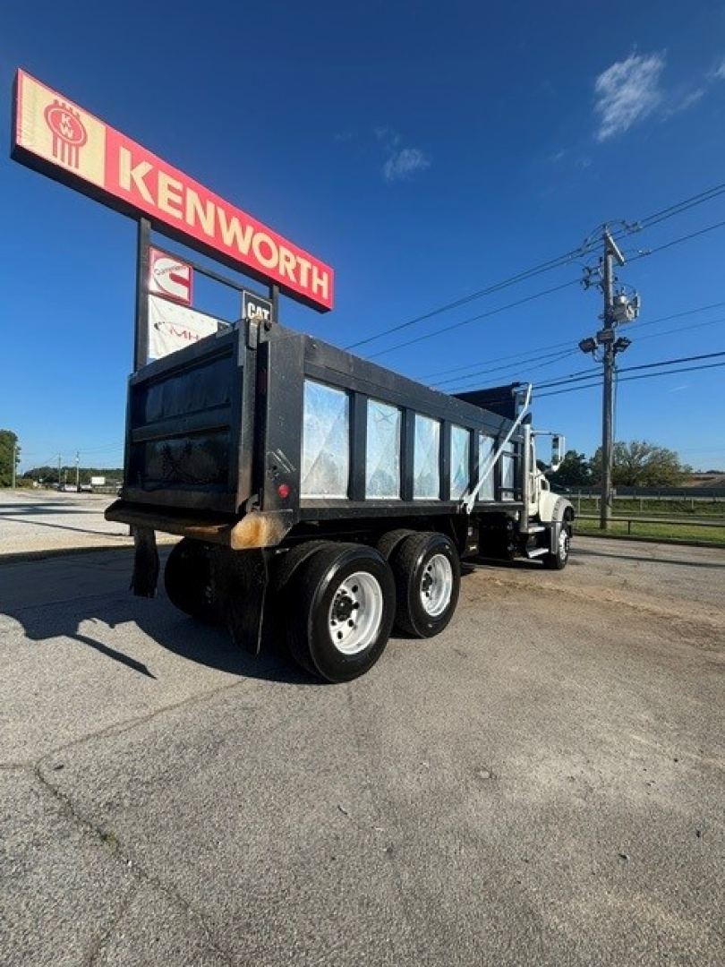 Dump truck side view showing Mack GRANITE dump body with dual rear axles under a black bed and white cab, Kenworth sign in background, clear sky.