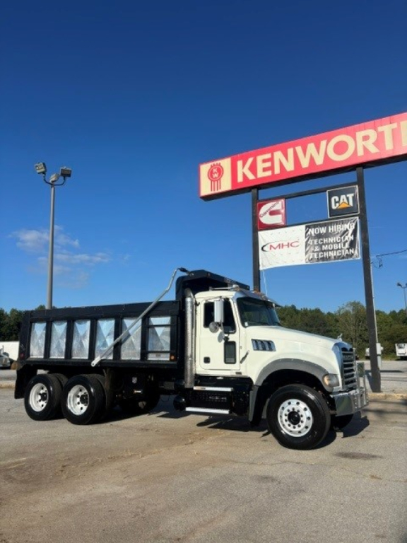 Truck in a Kenworth dealership lot showing a white Mack Granite dump truck with a black dump bed and three rear axles.