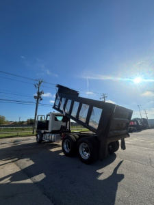 Dump truck in service showing a Mack GRANITE dump bed raised for dumping with white cab and black dump body.