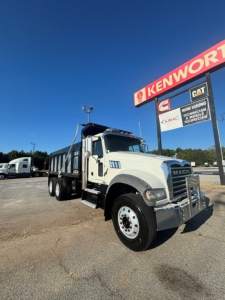 Mack Granite dump truck in a dealership lot, white cab with chrome bumper, large grille, dual rear axles, and dump body visible under bright blue sky.