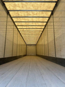 Van trailer interior cargo area with long rectangular ceiling lights, empty wooden floor, side walls, and rear door visible in distance.