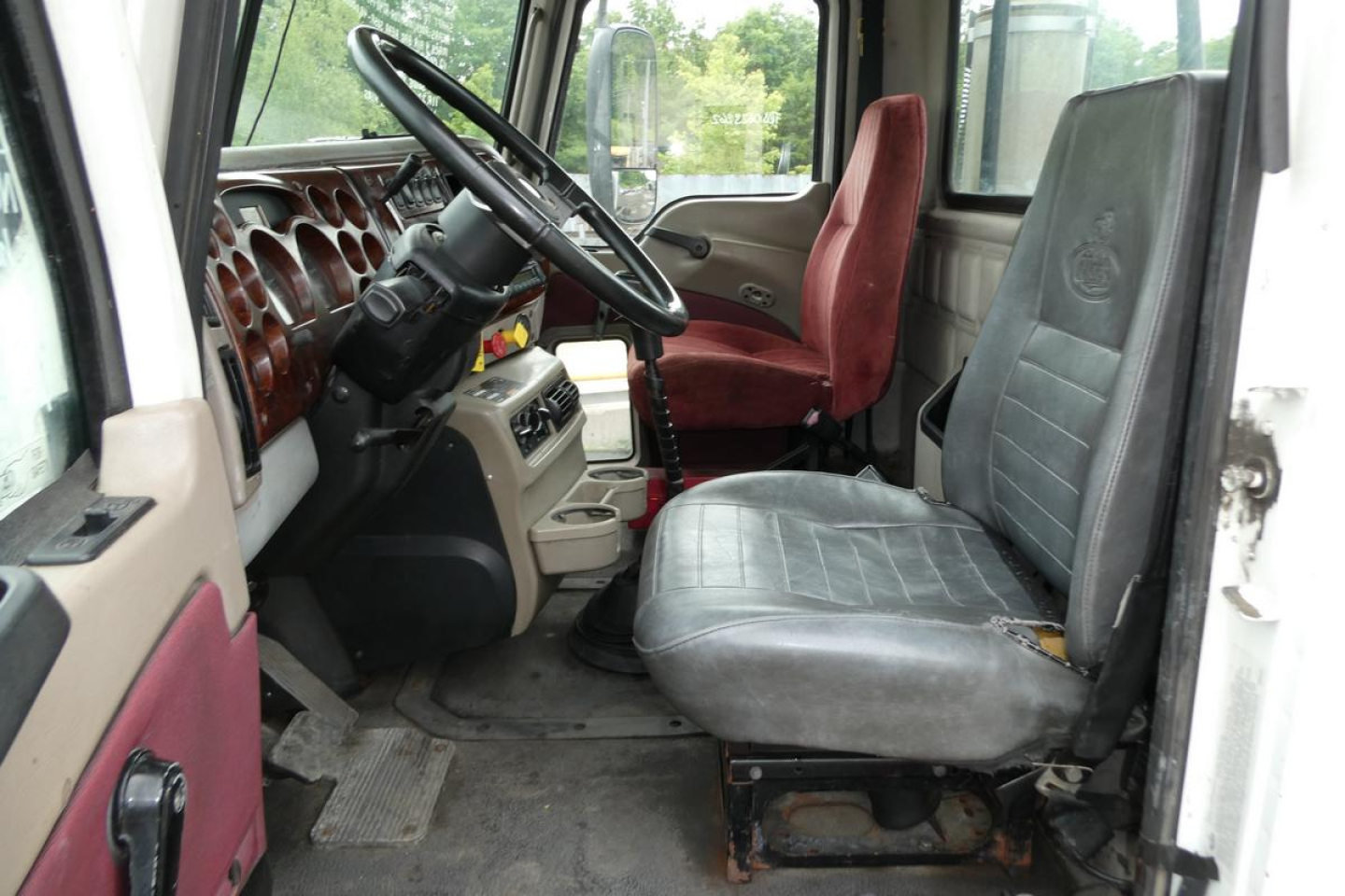Truck cab interior showing steering wheel, dashboard with numerous gauges, two front seats (driver and passenger) and a red passenger seat with wear.