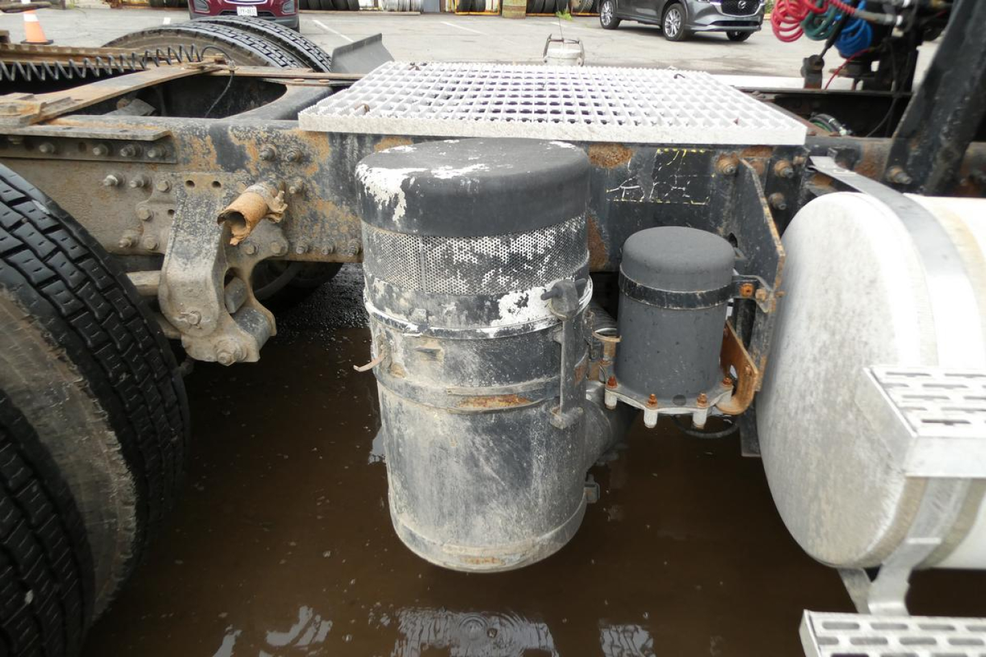 Truck tractor chassis showing cylindrical air reservoir and vertical black cylindrical canister with metal mounting, mud and grime on tanks, side steps, and frame.