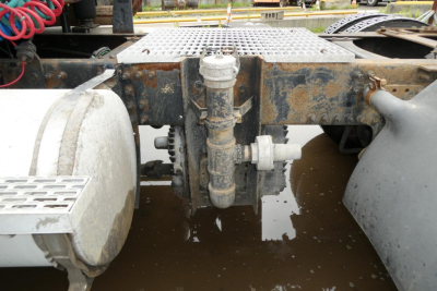 Truck chassis area showing air reservoir and plumbing fittings between drive wheels; mud, dirt, and water pooled on ground under central frame members.