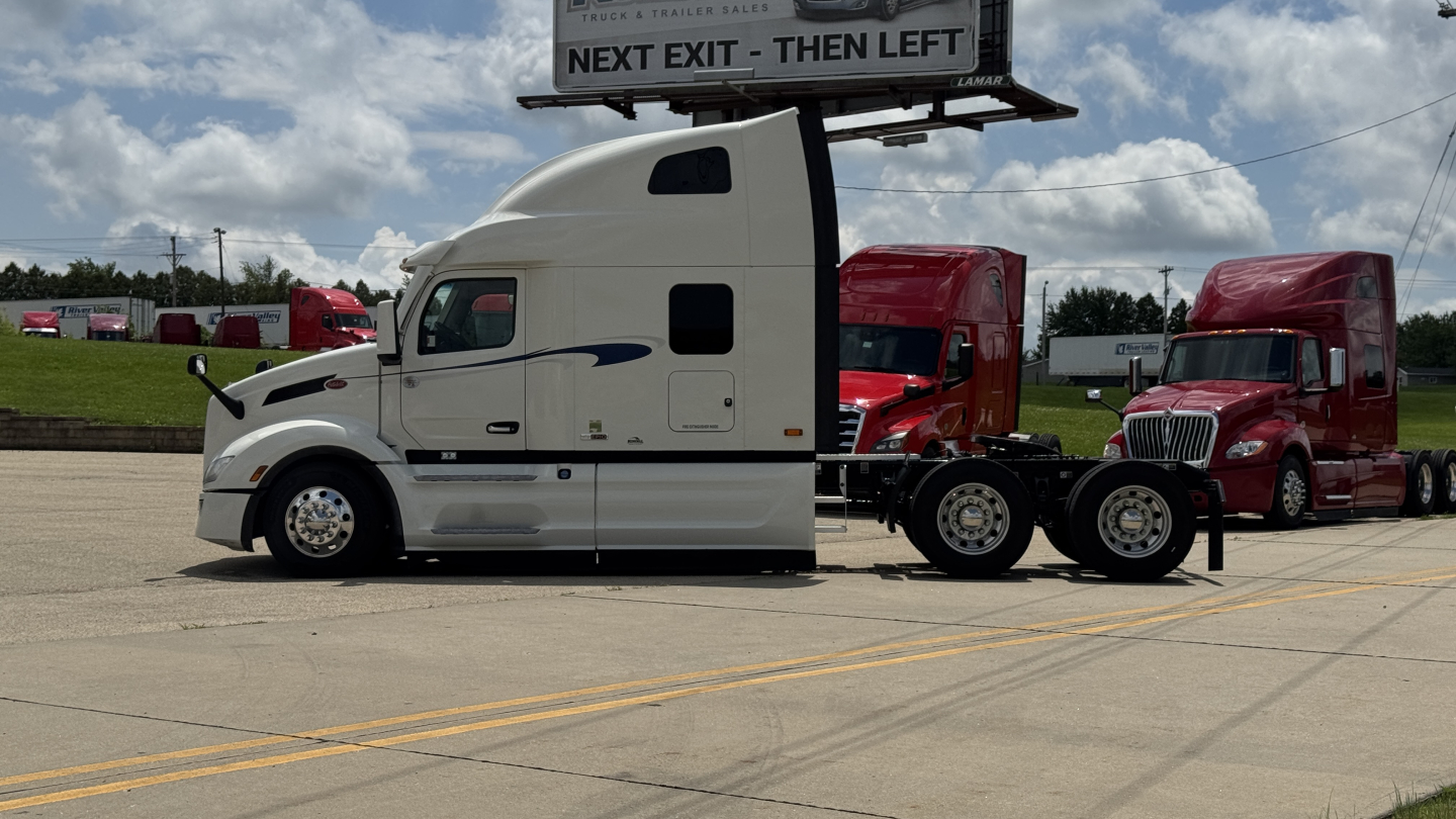 Peterbilt 579 tractor unit in a dealership lot, white long-nose cab with sleeper, black trim, side mirrors, and two red trucks in background.