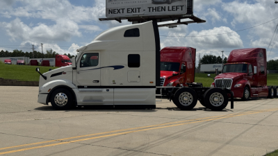 Peterbilt 579 tractor unit in a dealership lot, white long-nose cab with sleeper, black trim, side mirrors, and two red trucks in background.
