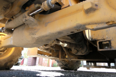Underbody view of a yellow heavy-duty lift truck showing a hydraulic cylinder, frame rails, and drivetrain components.