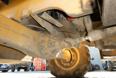 Truck lift undercarriage showing wheel and suspension components, yellow painted frame, hydraulic cylinder, axle joint, and steering linkages.