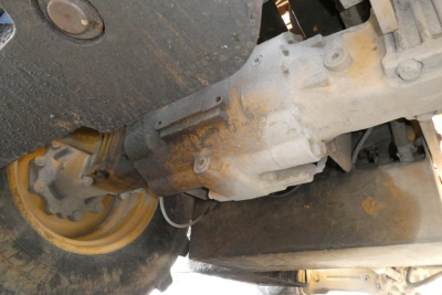 Underbody view of a truck lifting mechanism showing a gear housing and wheel with a yellow rim; rusty grime on metal surfaces and exposed hydraulic components.