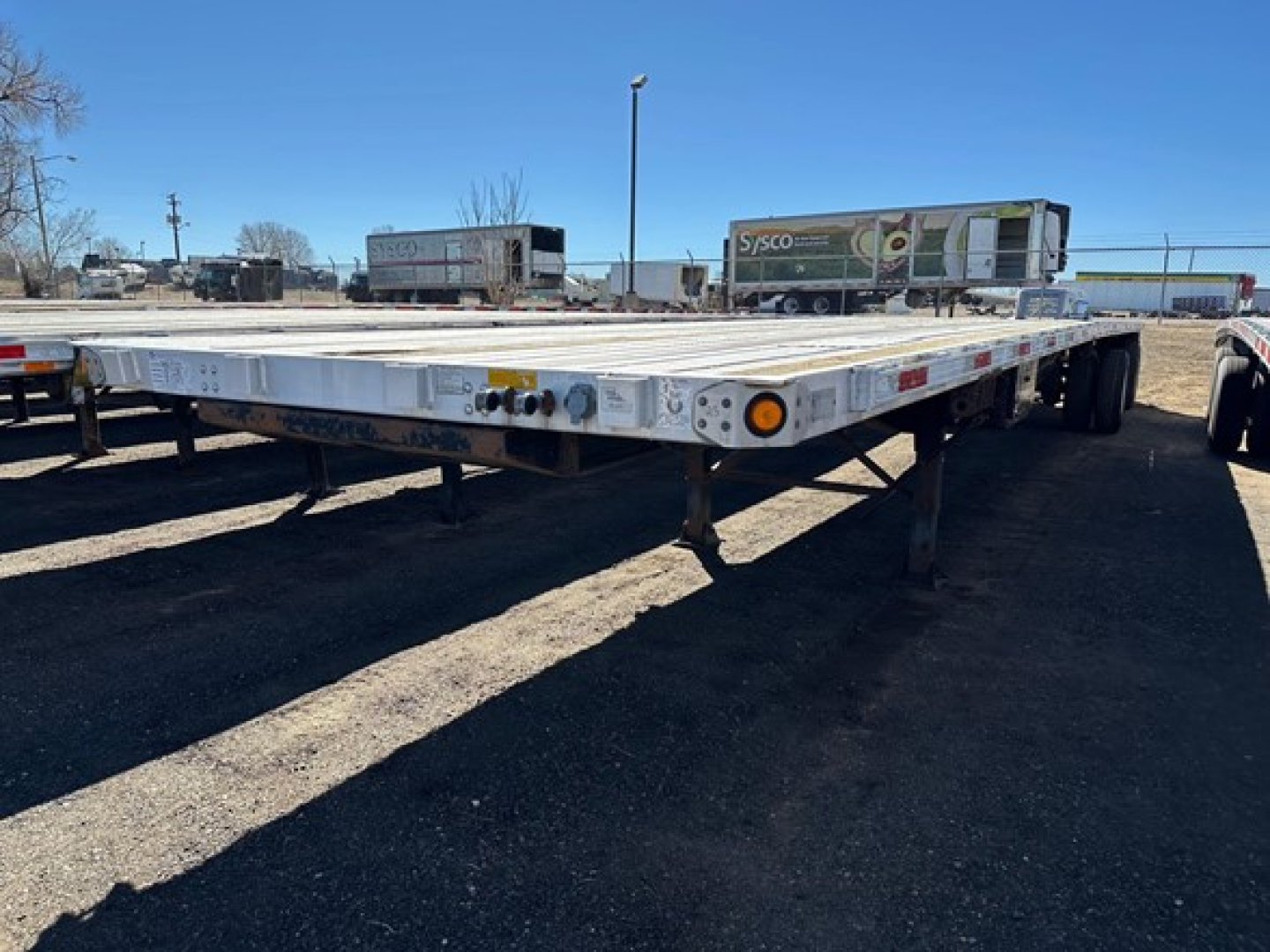 Flatbed trailer with aluminum deck and dual rear axles in a storage yard.