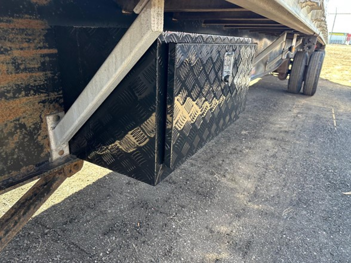 Flatbed trailer side view showing underbody diamond-plate toolbox and hardware with dual rear axles.