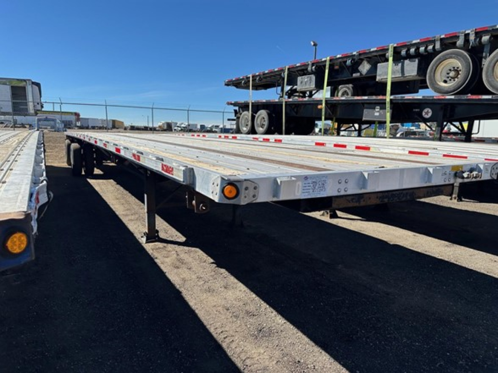Flatbed trailer from side view showing aluminum deck with multiple stake pockets and reflectors, under a clear sky.