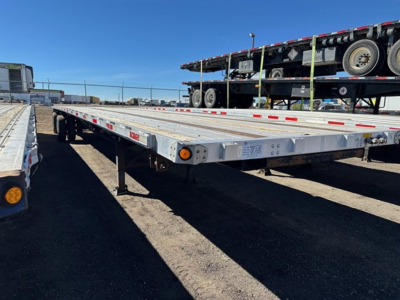 Flatbed trailer from a side angle showing aluminum decking and wheel rails with safety reflectors and a lifted secondary trailer in the background.