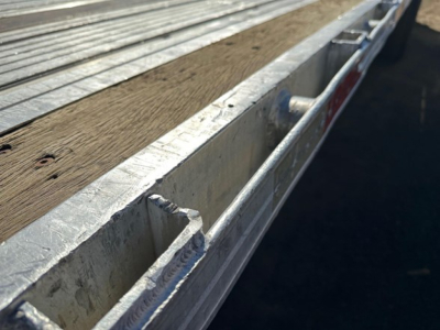 Aluminum utility flatbed trailer side rail and wooden deck visible, showing edge reinforcements and stake pocket with welds.