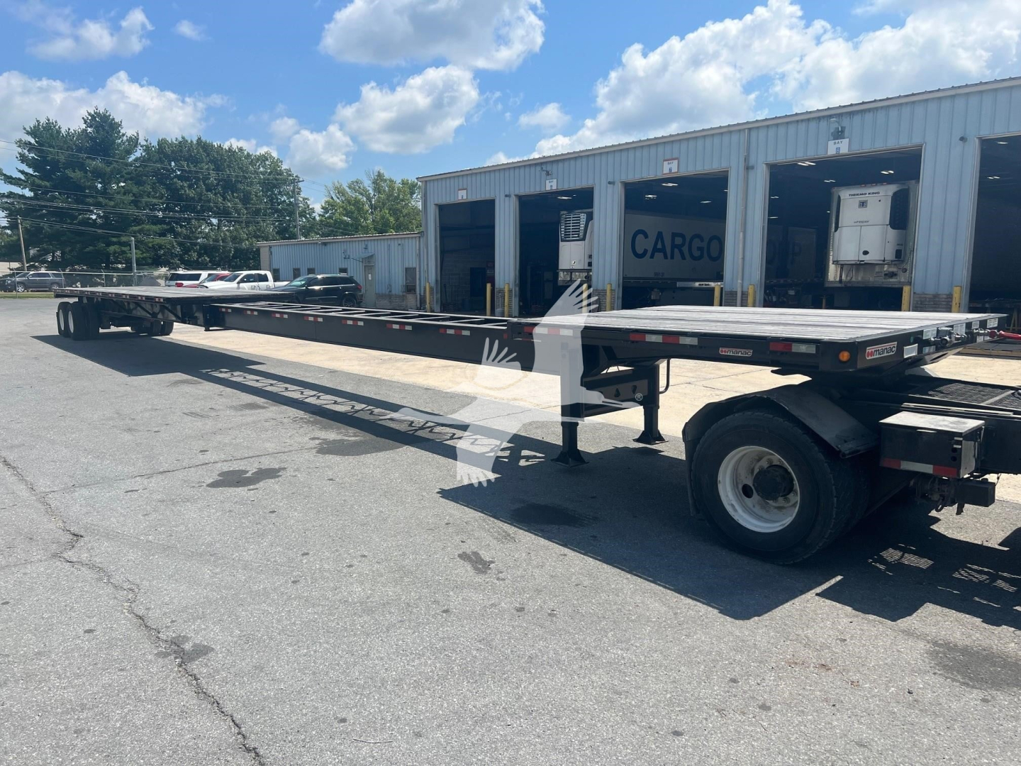 Manac air ride extendable flatbed trailer in a parking lot showing a long flatbed with rear wheels and support legs, warehouse in background.