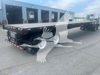 Flatbed trailer with extended air-ride feature, rear view showing tandem axles and reflective tape, Manac branding on side, loading dock in background.